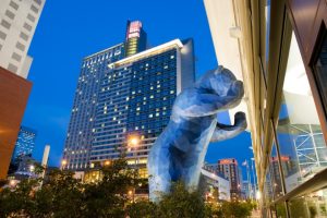 Groot blauw kunstwerk van een beer dat door een gevel naar binnen kijkt bij het Colorado Convention Center in Denver, met wolkenkrabbers op de achtergrond bij schemering