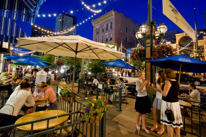 Mensen genieten van een terras met parasols en lichtjes op Larimer Square in Denver, met historische gebouwen op de achtergrond bij avond