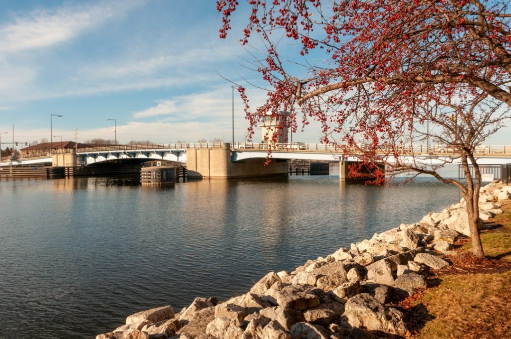 Betonnen ophaalbrug over kalm water met een stenen kade op de voorgrond en een boom met rode besjes