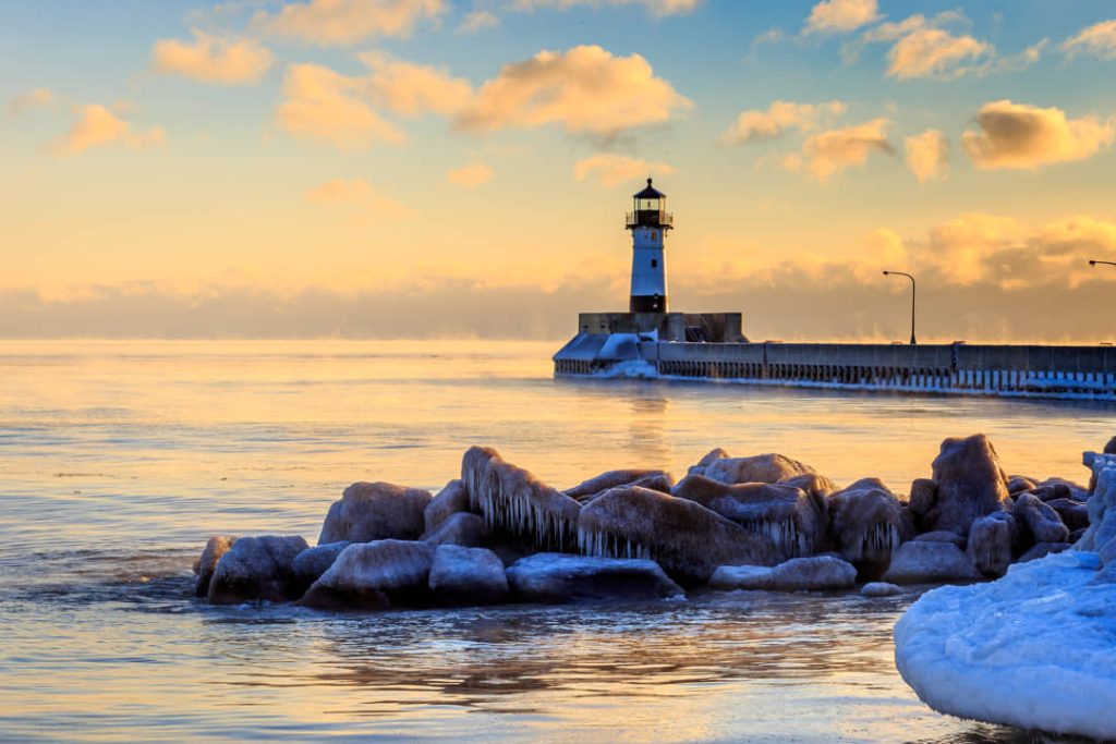 Witte vuurtoren aan het einde van een pier in Duluth, met bevroren rotsen op de voorgrond bij een gouden zonsondergang