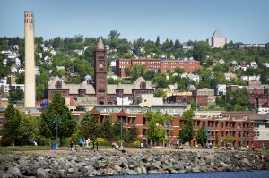 Skyline van Duluth met een oude schoorsteen, een klokkentoren en bakstenen gebouwen tegen een heuvel, gezien over een stenen dam