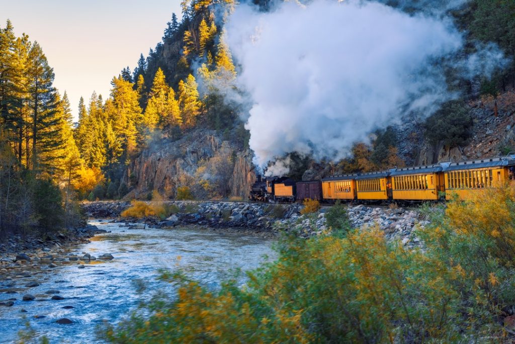Stoomlocomotief met oranje wagons stoot een grote rookwolk uit terwijl hij langs een rotsige rivier rijdt tussen herfstbomen in Colorado