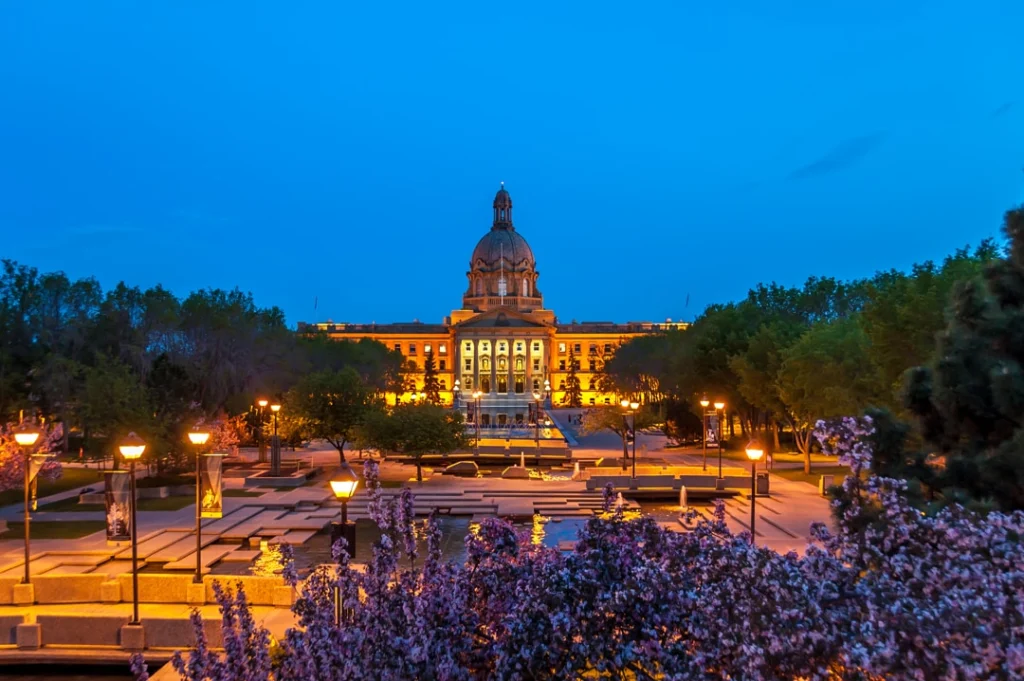 Het verlichte koepelgebouw van het Alberta Legislature in Edmonton straalt tegen de blauwe avondlucht, omringd door verlichte paden en bloeiende struiken