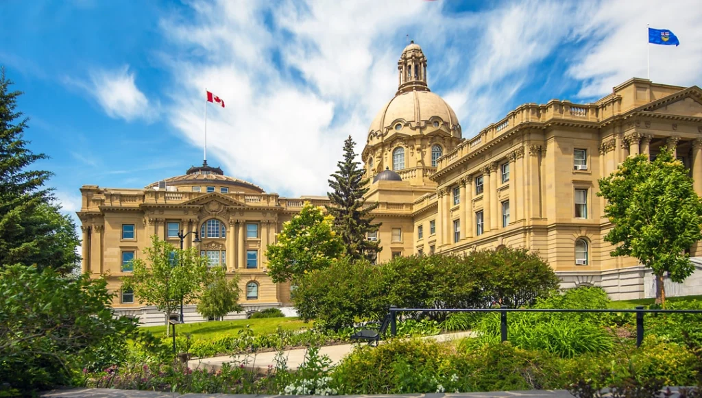 Zandkleurig regeringsgebouw Alberta Legislature in Edmonton met een koepeldak en wapperende vlaggen, omgeven door groen