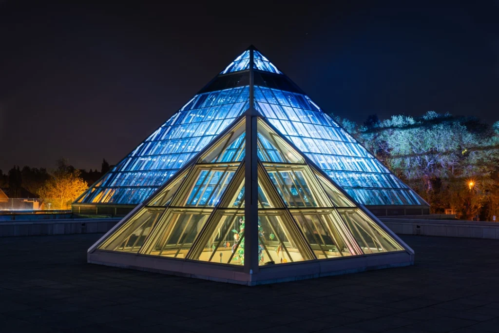 Een grote glazen piramide in Edmonton is 's nachts blauw verlicht tegen een donkere hemel met bomen op de achtergrond