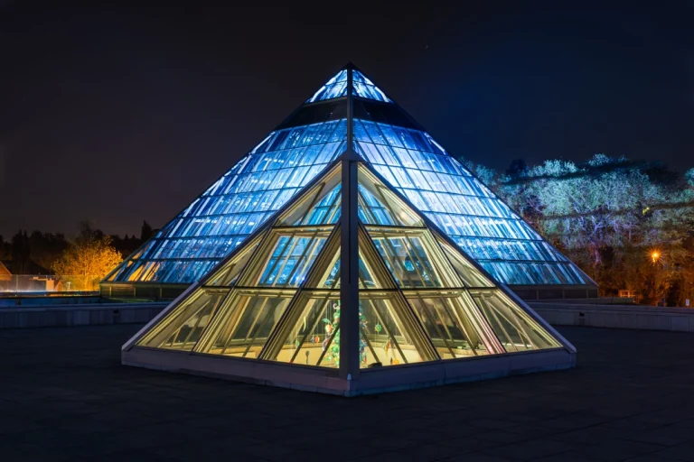 Een grote glazen piramide in Edmonton is 's nachts blauw verlicht tegen een donkere hemel met bomen op de achtergrond
