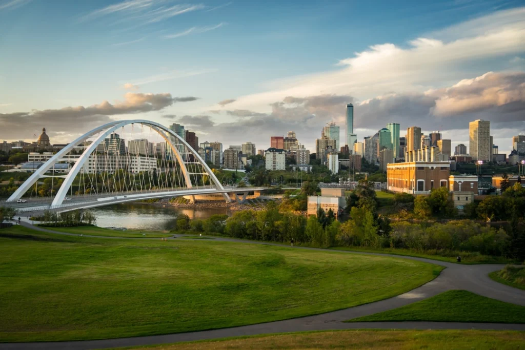 Witte boogbrug over de rivier met de skyline van Edmonton op de achtergrond, gezien vanaf een grasveld bij zonsondergang