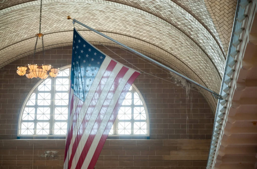 Grote Amerikaanse vlag hangend onder het gewelfde betegelde plafond van de registratiehal op Ellis Island met een kroonluchter en boograam