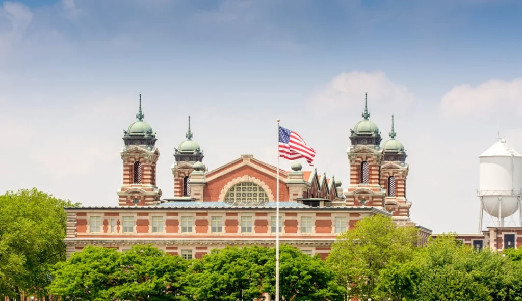 Rood bakstenen hoofdgebouw van Ellis Island met sierlijke torentjes, een wapperende Amerikaanse vlag en een watertoren onder een blauwe lucht
