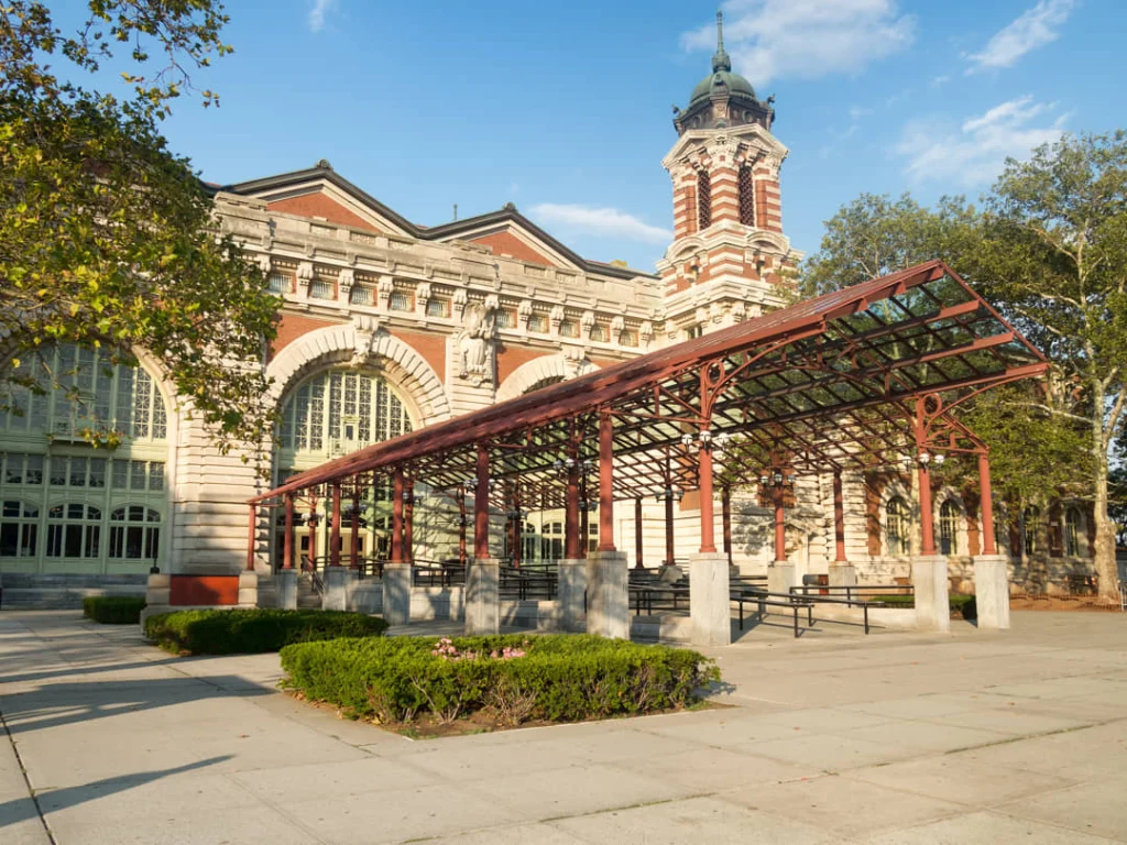 Rood bakstenen gebouw van Ellis Island met een sierlijke klokkentoren, grote rondboogramen en een moderne glazen overkapping bij de ingang