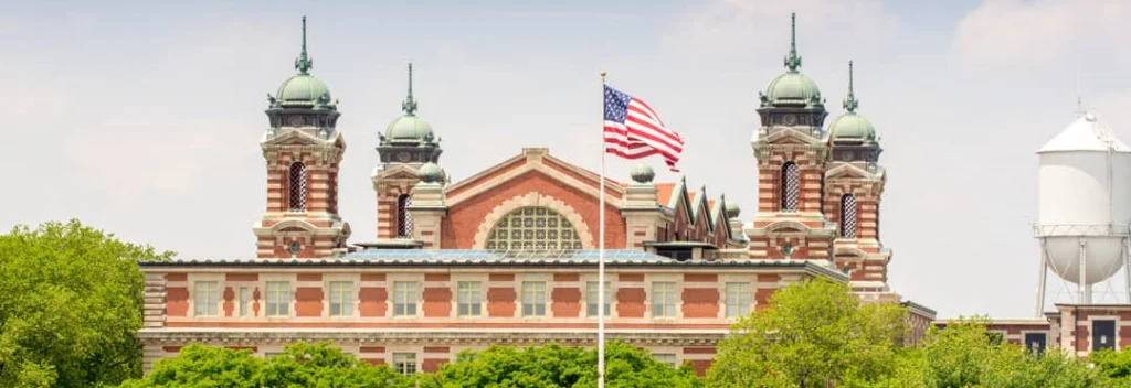 Close up van het rood bakstenen hoofdgebouw van Ellis Island met sierlijke koepeltorentjes en een wapperende Amerikaanse vlag ervoor