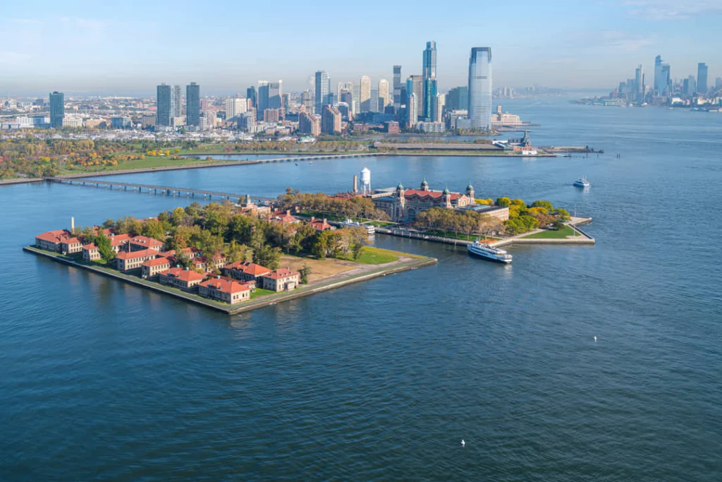 Luchtfoto van Ellis Island met zijn rode gebouwen en verbindingsbrug terwijl de wolkenkrabbers van Jersey City zichtbaar zijn