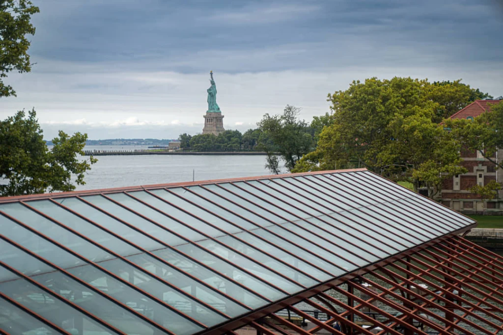Vrijheidsbeeld zichtbaar over het water tussen bomen gezien vanaf het glazen dak bij het hoofdgebouw van Ellis Island