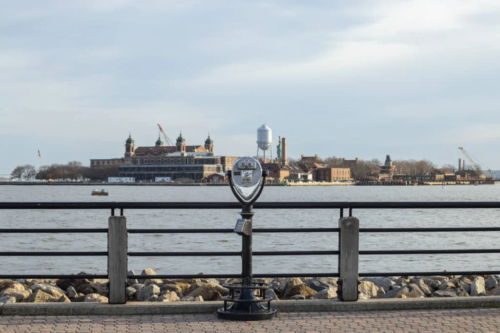 Muntverrekijker aan een hek langs het water met wazig op de achtergrond de gebouwen en watertoren van Ellis Island