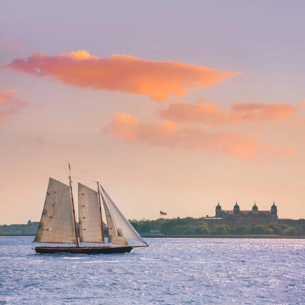 Zeilboot met witte zeilen die over het water vaart terwijl Ellis Island zichtbaar is onder een rozige avondlucht