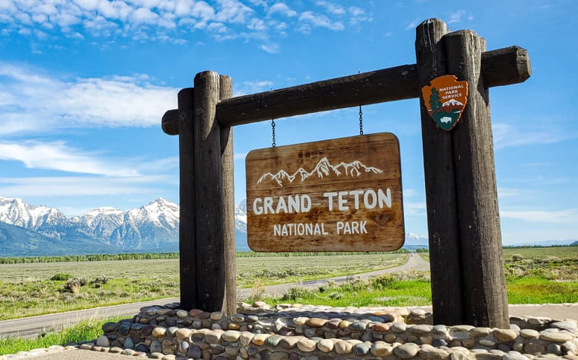 Een houten bord met opschrift Grand Teton National Park, met besneeuwde bergtoppen op de achtergrond