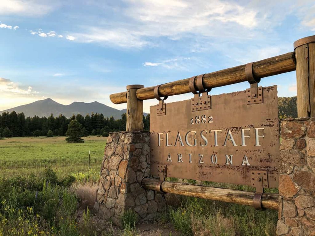 Houten en stenen welkomstbord met de tekst Flagstaff Arizona in een grasveld met bomen en bergen op de achtergrond