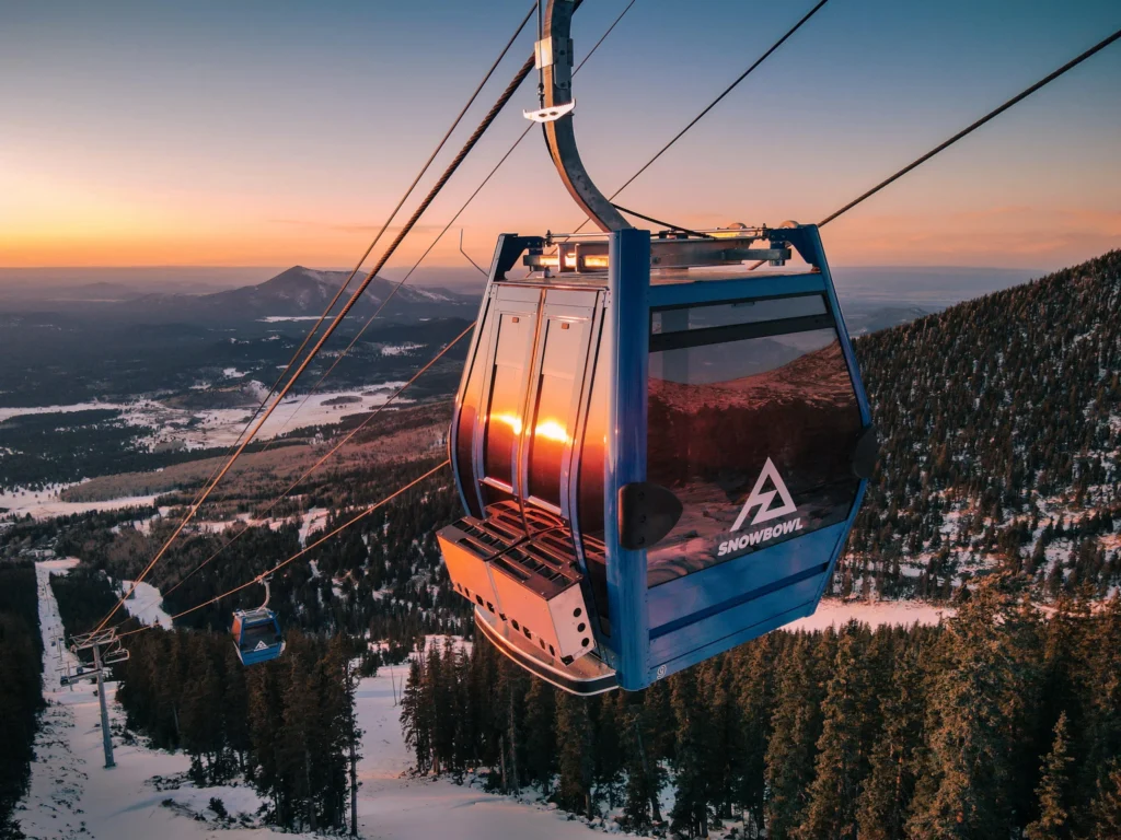 Blauwe gondelcabine van skigebied Arizona Snowbowl hangt boven besneeuwde pistes en bos terwijl de zon achter de heuvels ondergaat