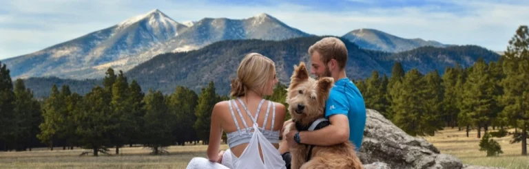 Man en vrouw zitten met hun hond op een rots in een grasveld met besneeuwde bergtoppen en dennenbos op de achtergrond