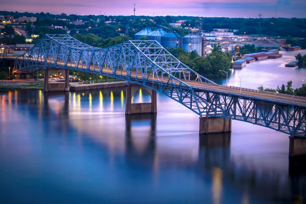 Blauwe stalen boogbrug over de Tennessee River met verlichte pylonen en een sleepboot met duwbakken bij avondschemering