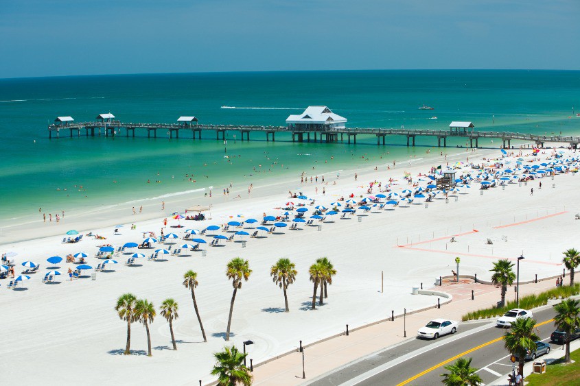 Luchtfoto van Clearwater Beach in Florida met blauwe strandparasols, palmbomen en de lange pier 60 die de zee in steekt