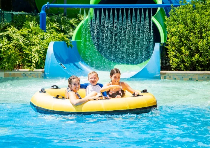 Moeder en twee kinderen drijven lachend in een gele band onder een groene waterglijbaan in een waterpark