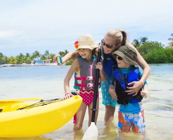 Moeder met twee kinderen in zwemvesten staat naast een gele kajak in ondiep water op een strand in Florida