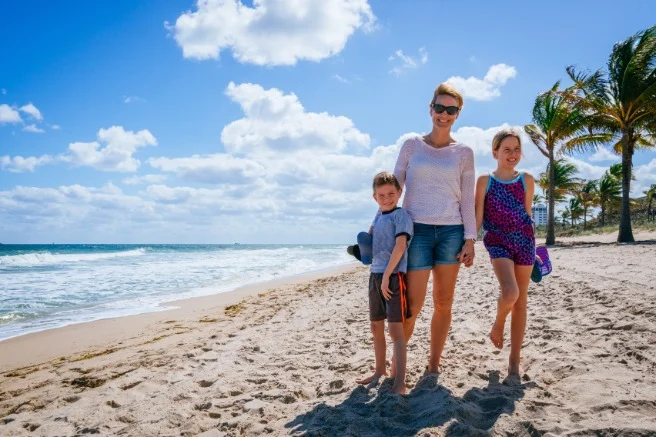 Moeder met zoon en dochter staan hand in hand op een zandstrand met golven en palmbomen op de achtergrond in Florida