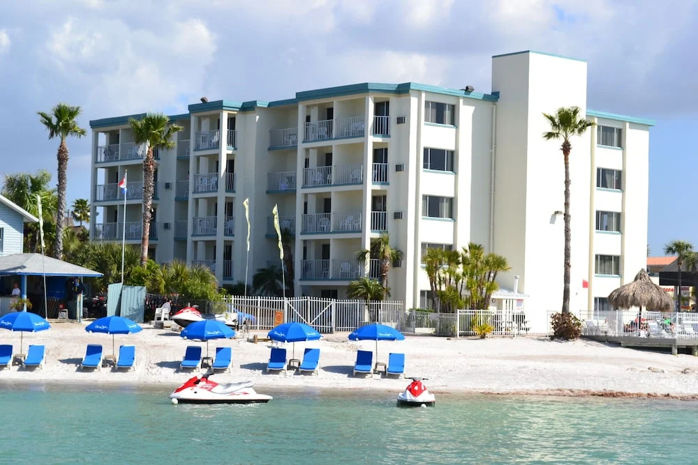 Wit hotelgebouw met balkons aan een zandstrand in St. Pete Clearwater, met blauwe strandstoelen, parasols en jetski's op het water