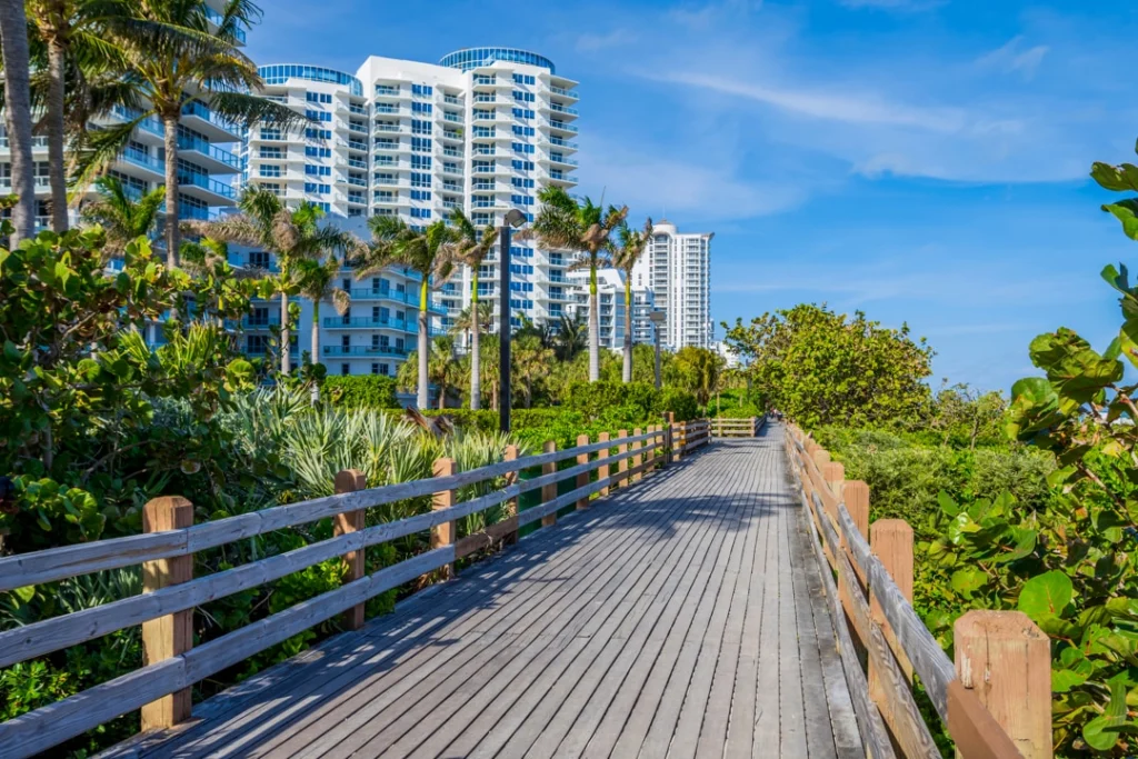 Houten wandelpad tussen struiken met hoge appartementengebouwen en palmbomen op de achtergrond onder een blauwe hemel in Florida