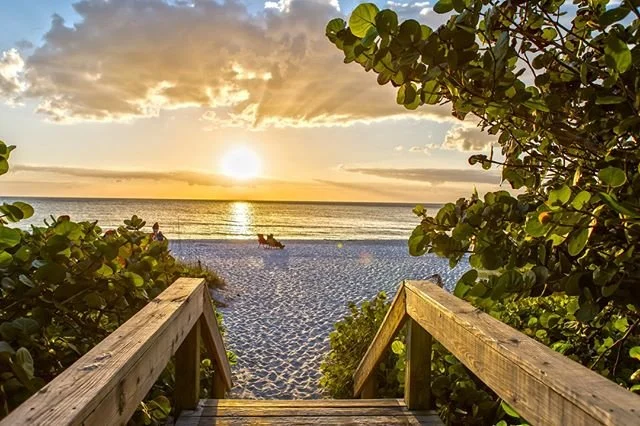Een houten trap tussen groene struiken voert naar een wit zandstrand in Florida terwijl de zon ondergaat boven zee