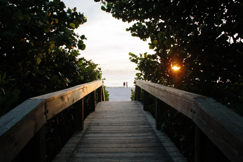 Een houten vlonder omgeven door groene struiken voert naar een zonnig strand bij Naples in Florida