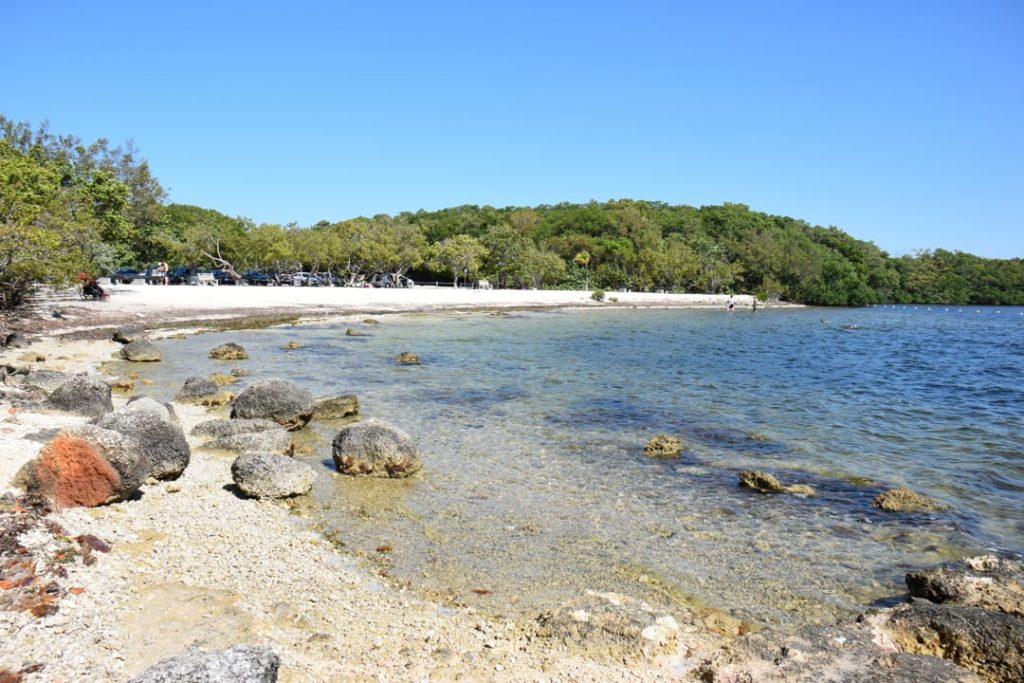 Kalme baai met rotsachtige oever, wit zand en mangrovebos aan de overkant onder een strakblauwe lucht