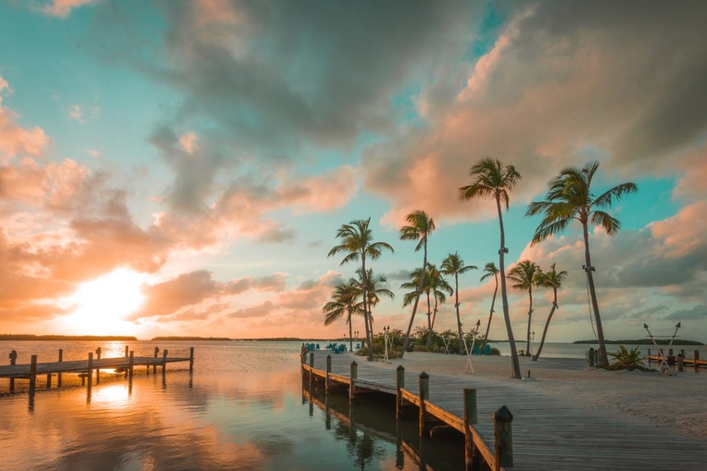 Houten steiger loopt de zee in tussen palmbomen onder een oranje en turquoise wolkenlucht bij zonsondergang