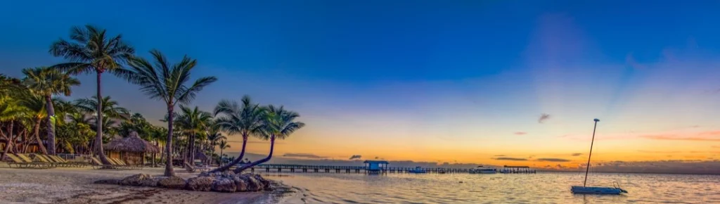 Ondiepe zee met een zeilboot en een steiger bij zonsondergang, met palmbomen en strandhutjes op een strand in de Florida Keys