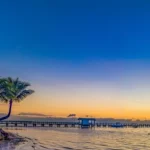 Ondiepe zee met een zeilboot en een steiger bij zonsondergang, met palmbomen en strandhutjes op een strand in de Florida Keys