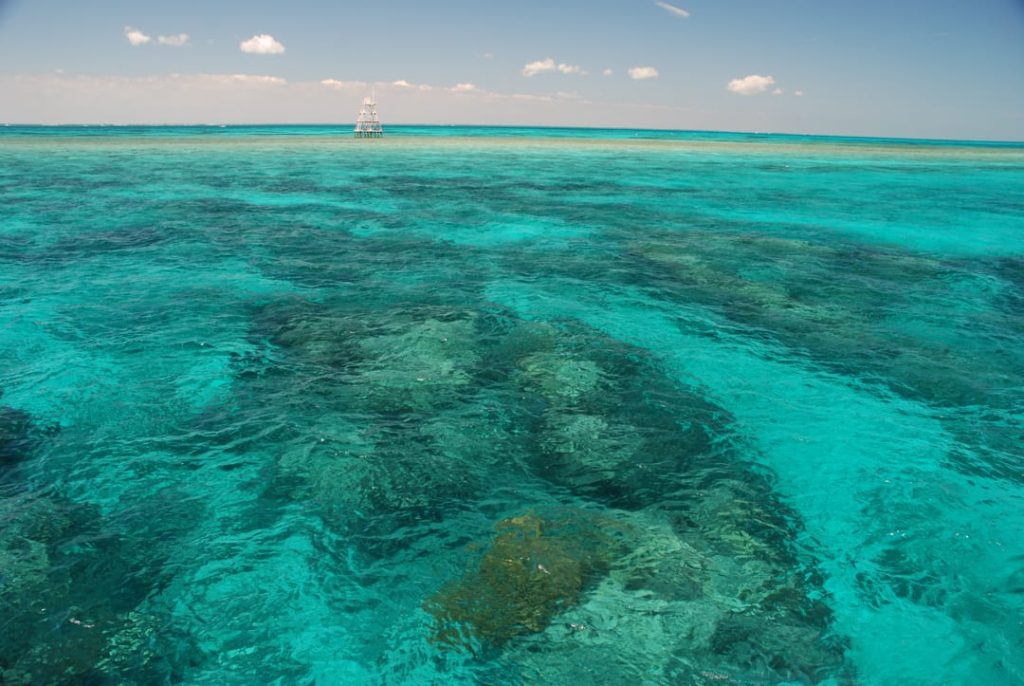 Helder turquoise water boven een uitgestrekt koraalrif met een metalen baken aan de horizon