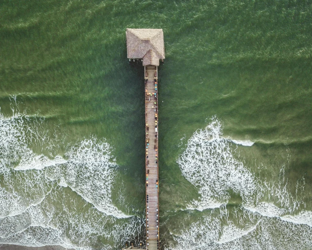 Luchtfoto van een lange pier met een gebouw met rieten dak aan het uiteinde, omringd door groenblauw water en golven in Florida