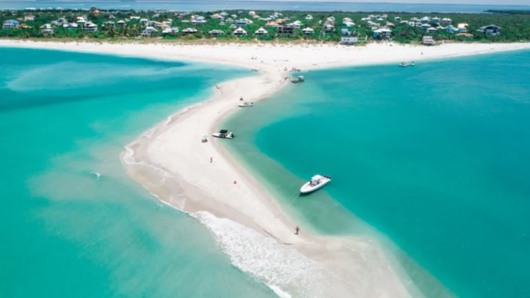 Luchtfoto van een smalle witte zandbank in Florida die uitsteekt in turquoise zee, met aangemeerde boten en een kustdorp op de achtergrond