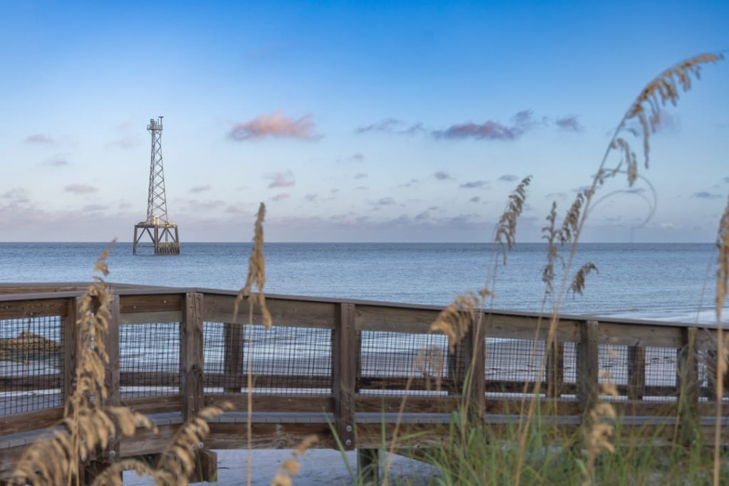 Metalen navigatiebaken in kalme zee gezien vanaf een houten boardwalk met strandgras op de voorgrond