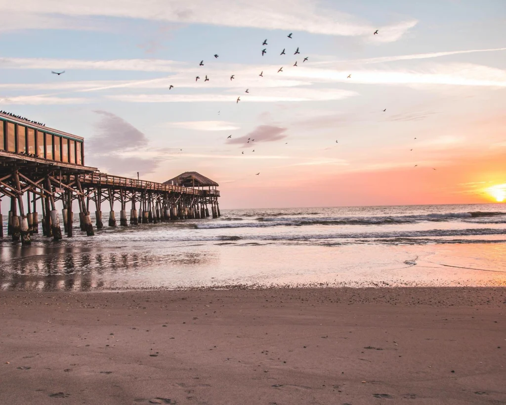 Houten pier op palen bij zonsondergang met een zwerm vogels boven het strand en de oceaan in Florida