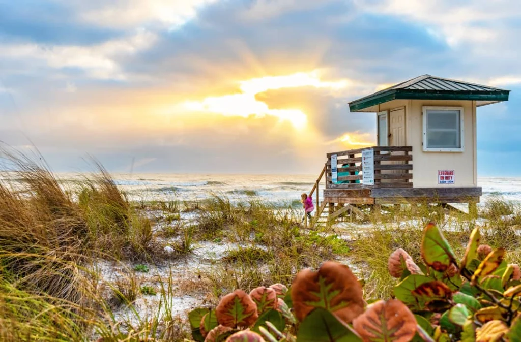 Wit reddingspostje met groen dak tussen strandgras, met een kind op de trap bij zonsopkomst boven zee
