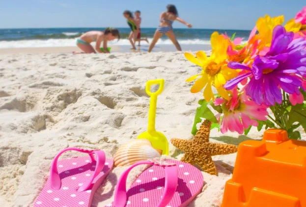 Roze slippers, een schelp, een zeester en een schepje liggen op het zand terwijl kinderen op de achtergrond spelen in zee