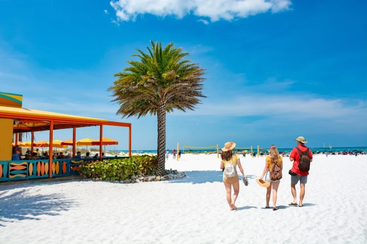 Ouders en kinderen met strandtassen lopen over wit zand langs een palmboom en een kleurrijke strandbar met oranje parasols in Florida
