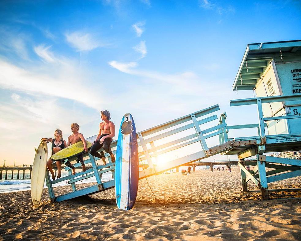 Drie surfers met surfplanken zitten op de leuning van een blauwe reddingspost op het strand terwijl de zon ondergaat