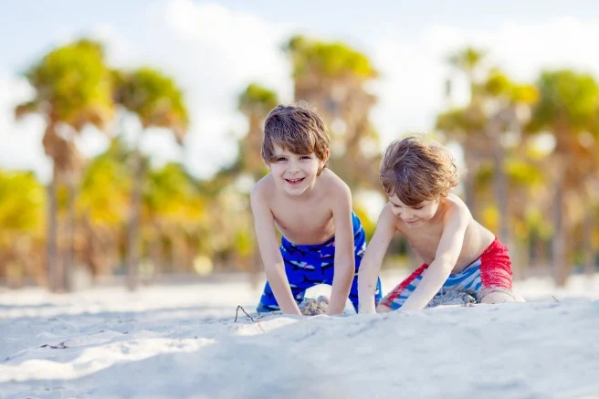 Twee lachende jongens in zwembroek graven met hun handen in wit zand met een wazige achtergrond van palmbomen op een zonnig strand