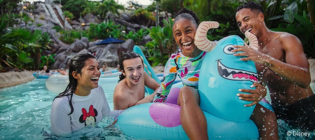 Vier vrienden lachen in het water terwijl een van hen op een grote blauwe opblaasfiguur zit in een Disney waterpark