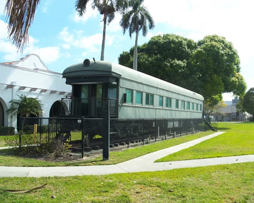 Groene historische spoorwegwagon achter een hek naast een wit gebouw met palmbomen op een museumterrein
