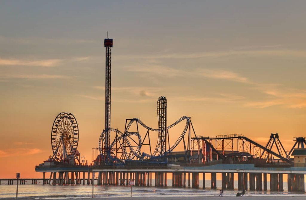 Silhouet van een pretpark met reuzenrad en achtbaan op een pier boven zee bij een oranje zonsondergang in Galveston