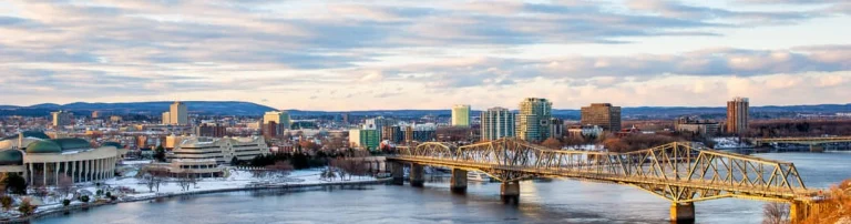 Een lange metalen brug over de rivier met op de achtergrond de skyline van Gatineau in het avondlicht